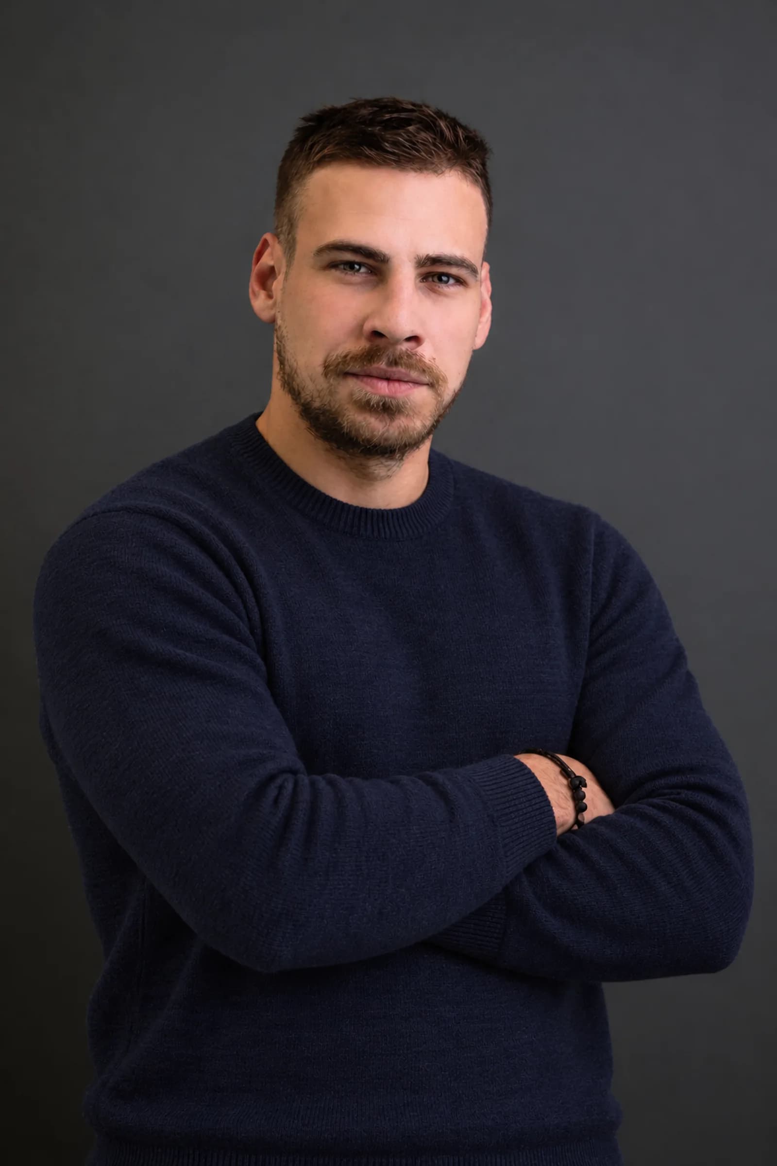 Damir Kozaragić, arms folded, in a navy sweater against a dark grey studio background.