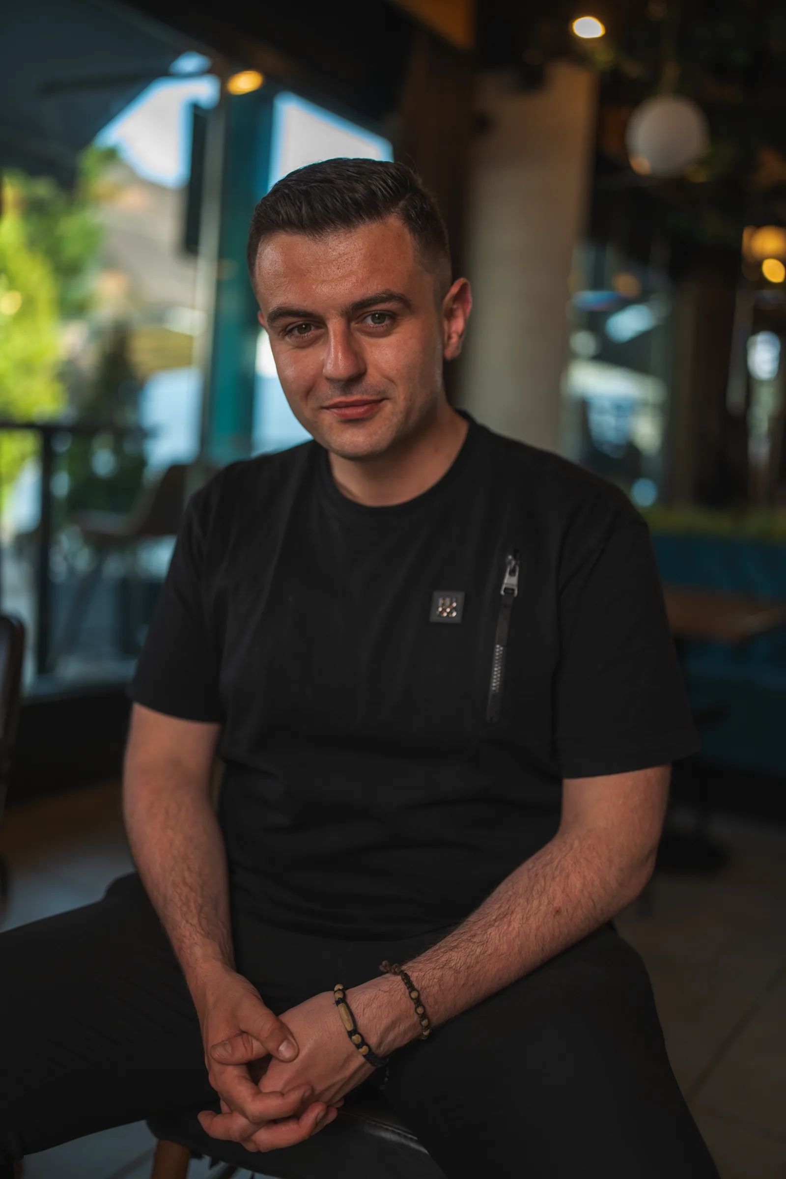 Tihomir Hrvacevic, seated with his hands clasped, in a black T-shirt, in a café with a window behind him.