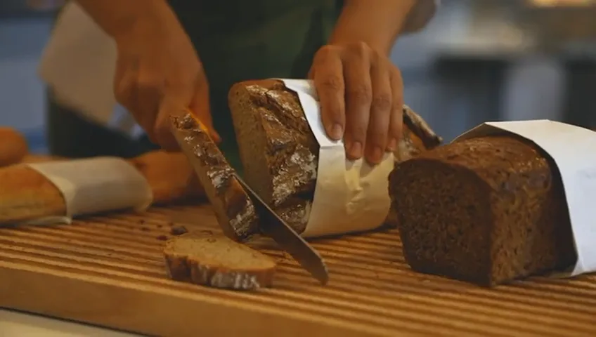 Hands slicing a dark loaf on a wooden board at the breakfast buffet.