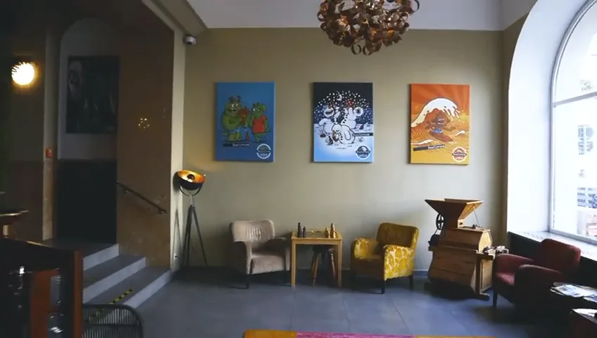 The hotel lounge: armchairs and a small chess table under three bright posters and a tall arched window.