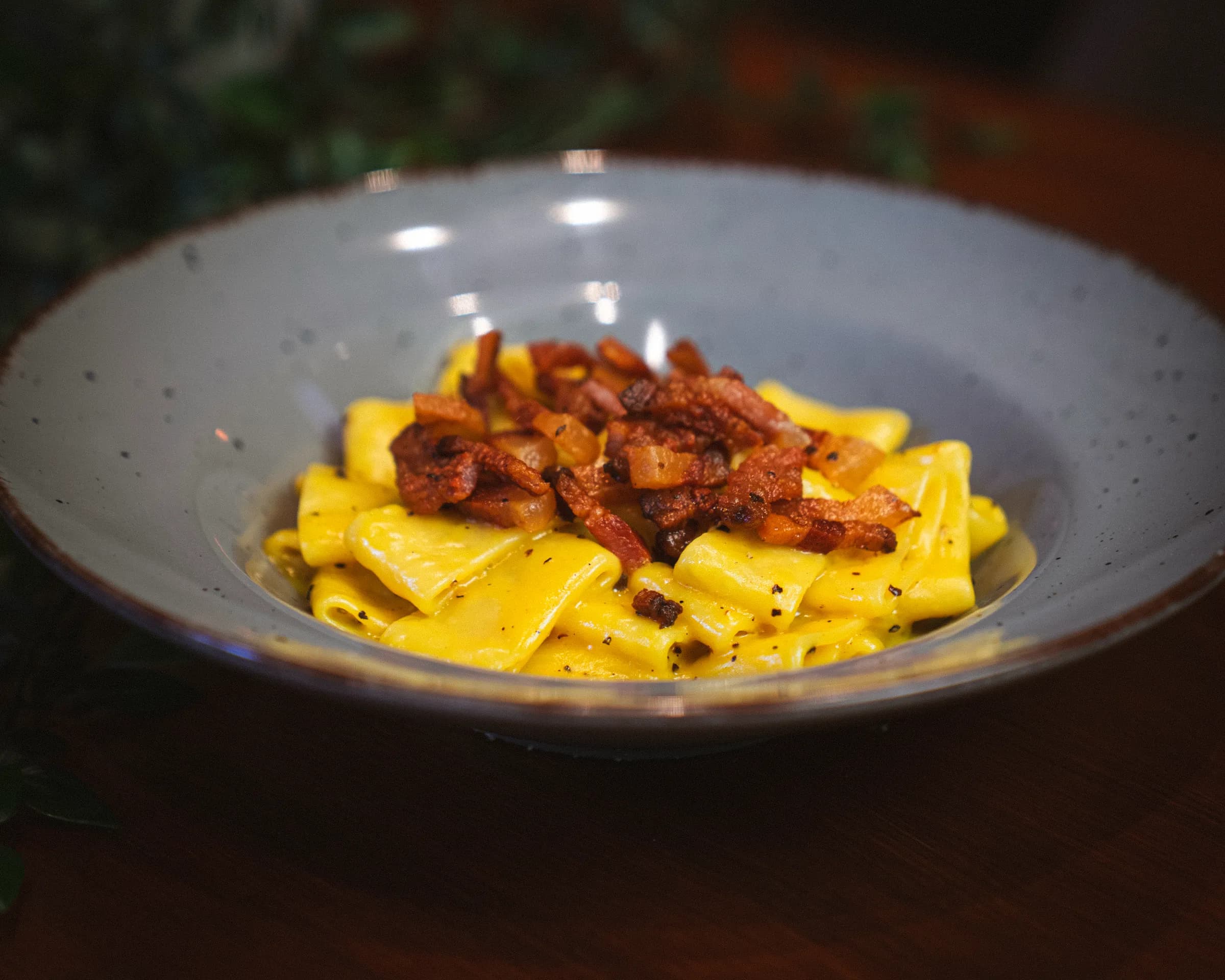 Wide pasta in a glossy yellow sauce, topped with crisp pieces of cured pork, in a grey speckled bowl.