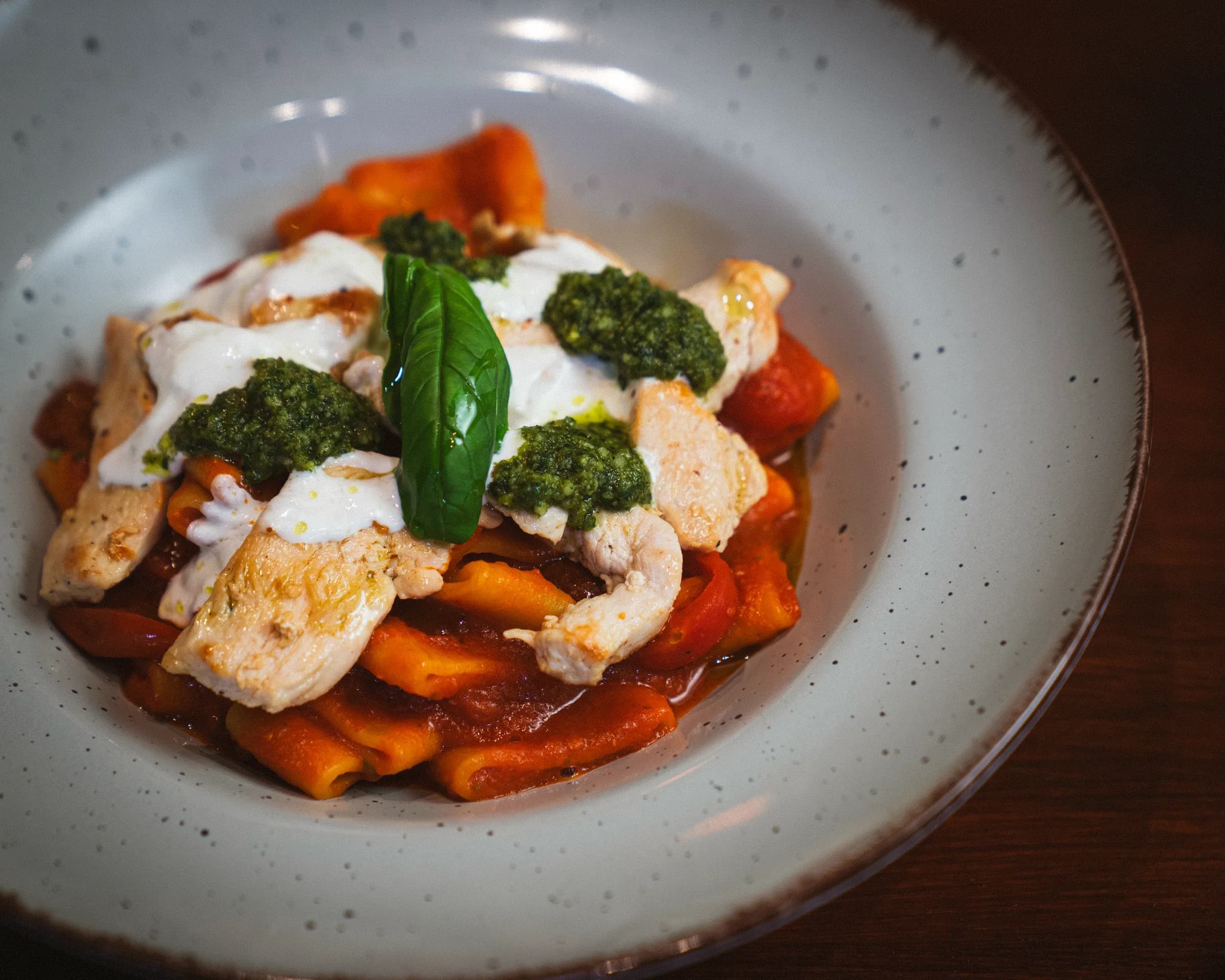 Short pasta in tomato sauce with pieces of chicken, cream, spoonfuls of pesto and a basil sprig, in a speckled bowl.