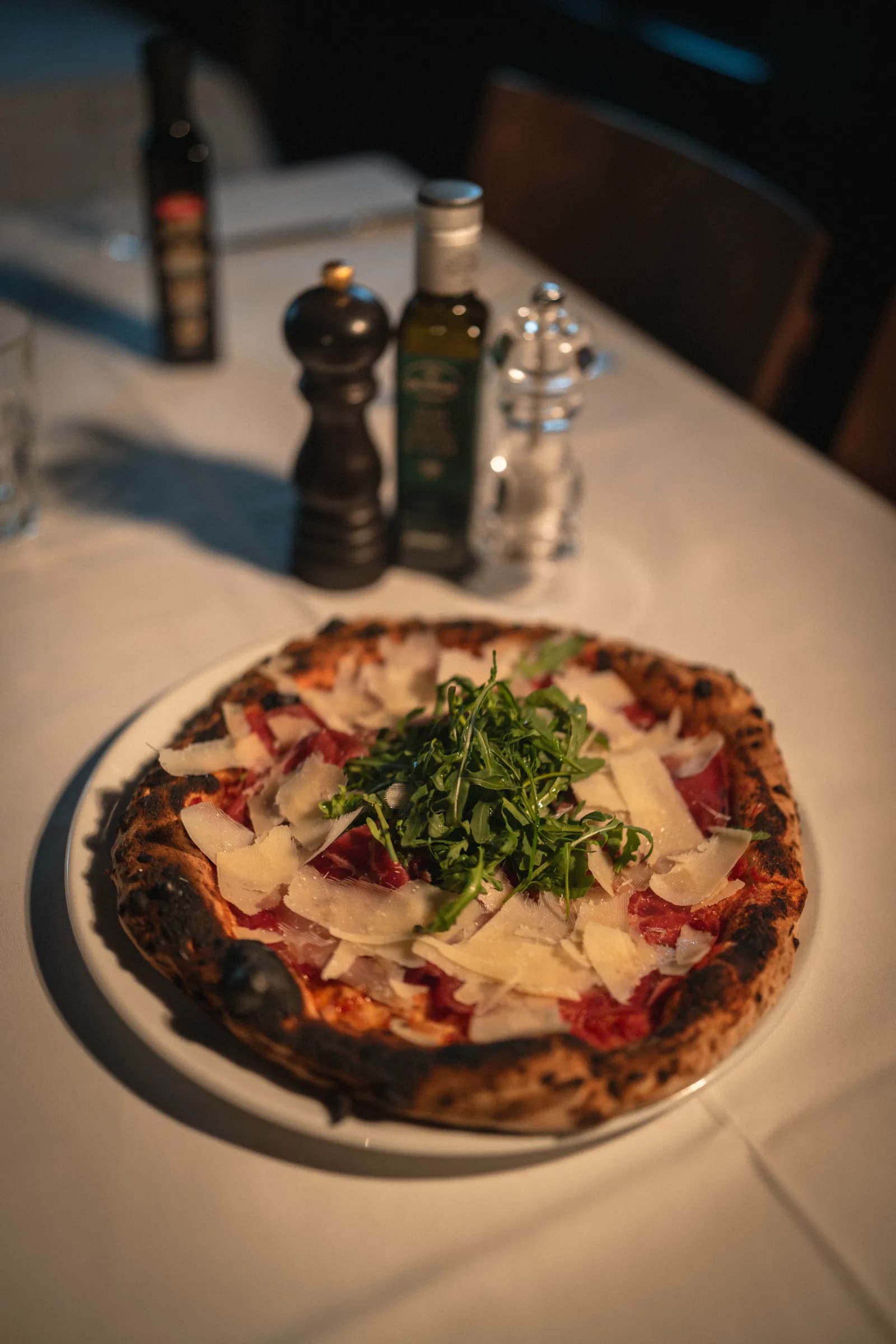 A pizza with bresaola, rocket and shaved parmesan on a white tablecloth, olive oil and a pepper mill behind it.