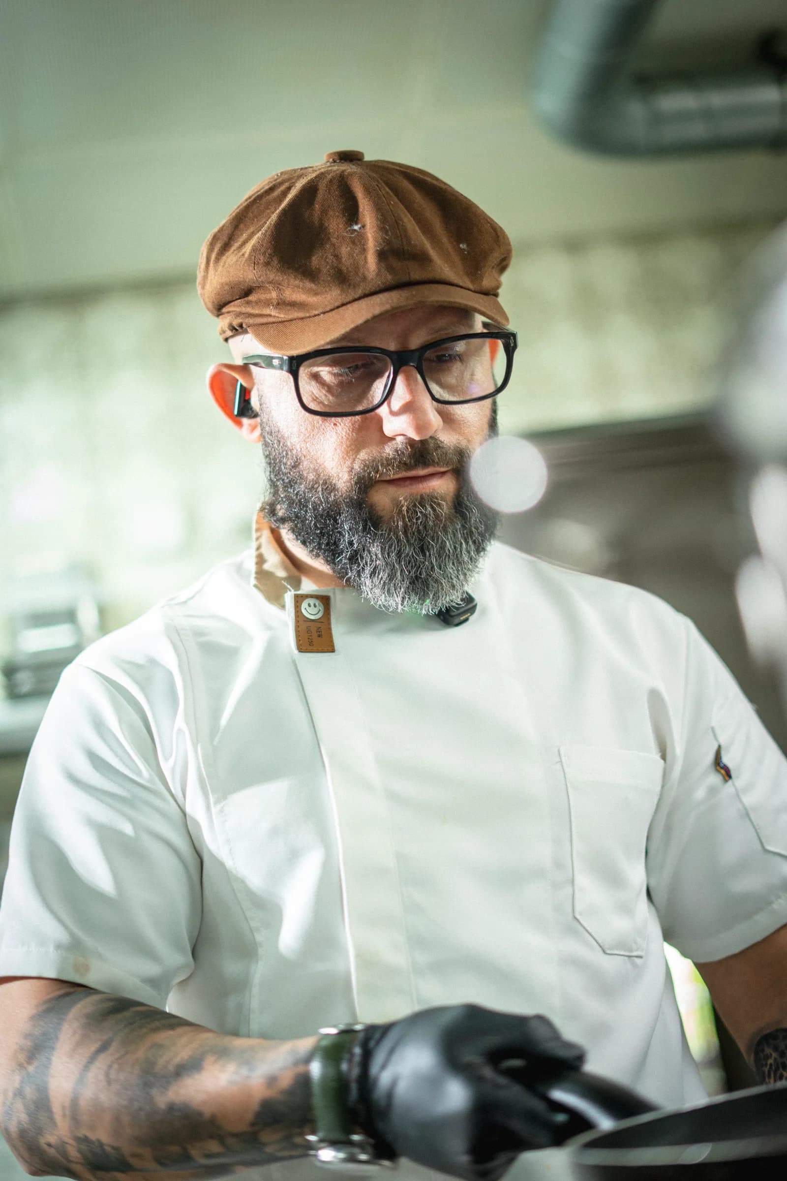 A chef in a white jacket, flat cap and glasses, looking down at his work in the kitchen.
