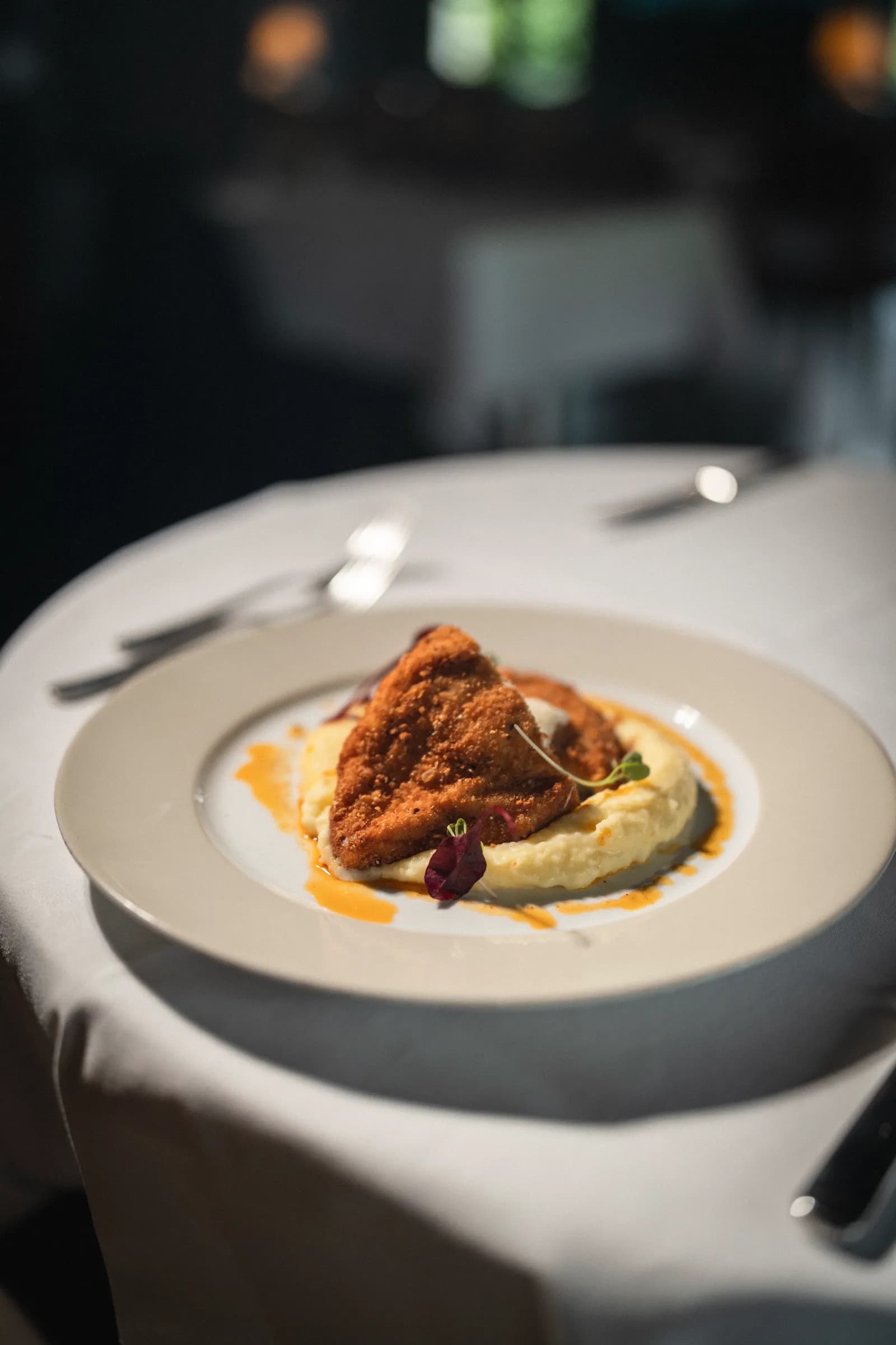 A breaded cutlet on potato purée with an orange sauce, on a white-clothed table in a dim dining room.