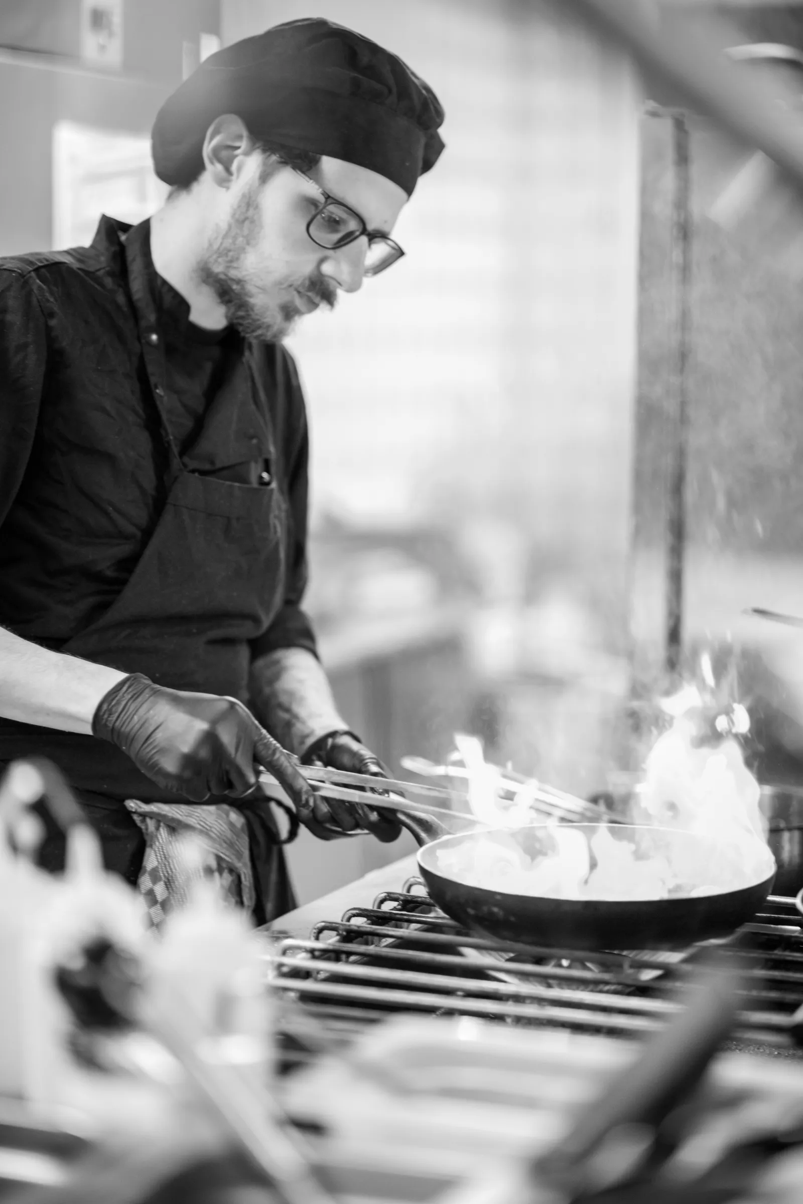 Black and white: a cook tossing a pan over a gas burner, flames rising from the pan.