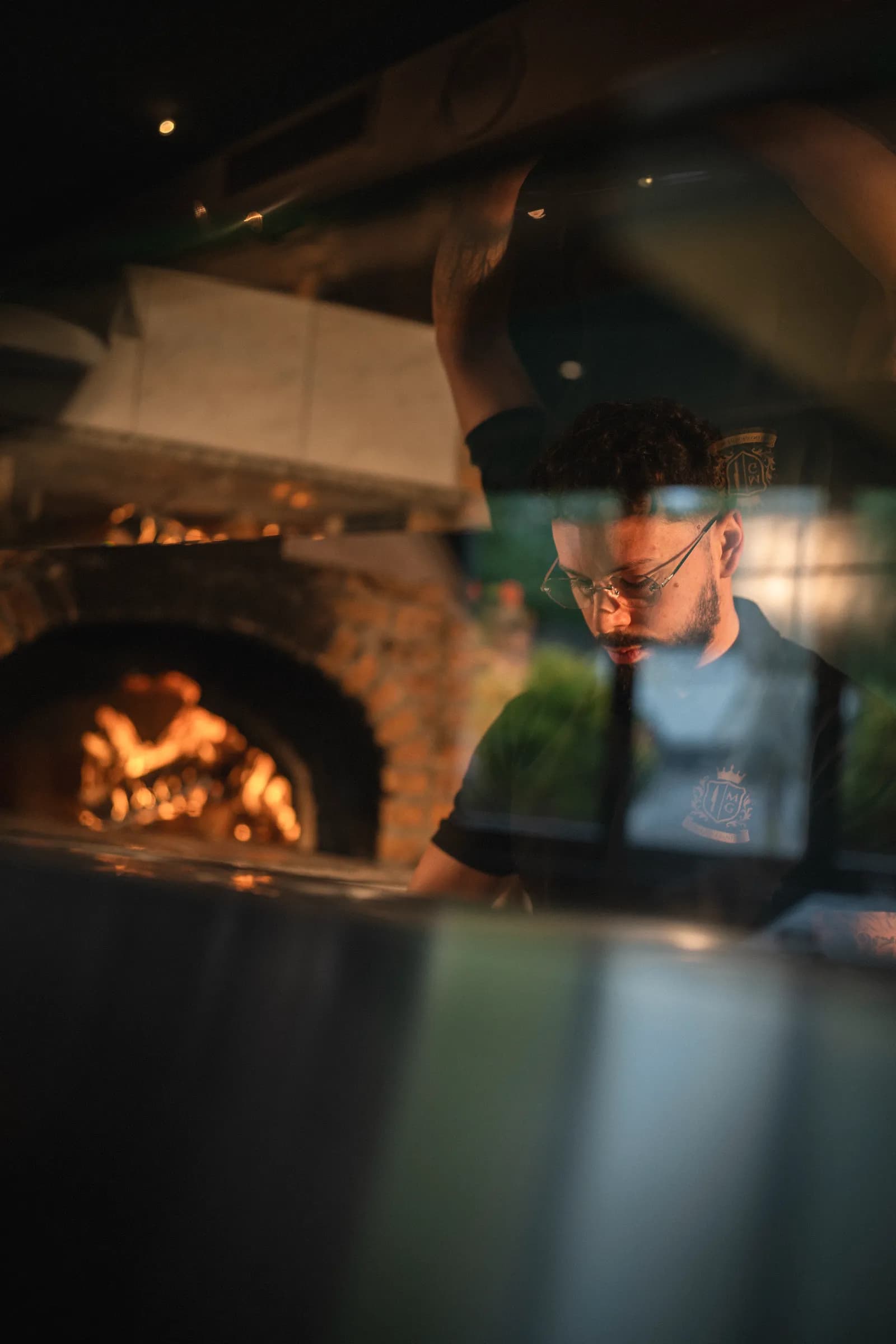 A pizzaiolo behind the counter, head down, the wood-fired oven burning behind him.