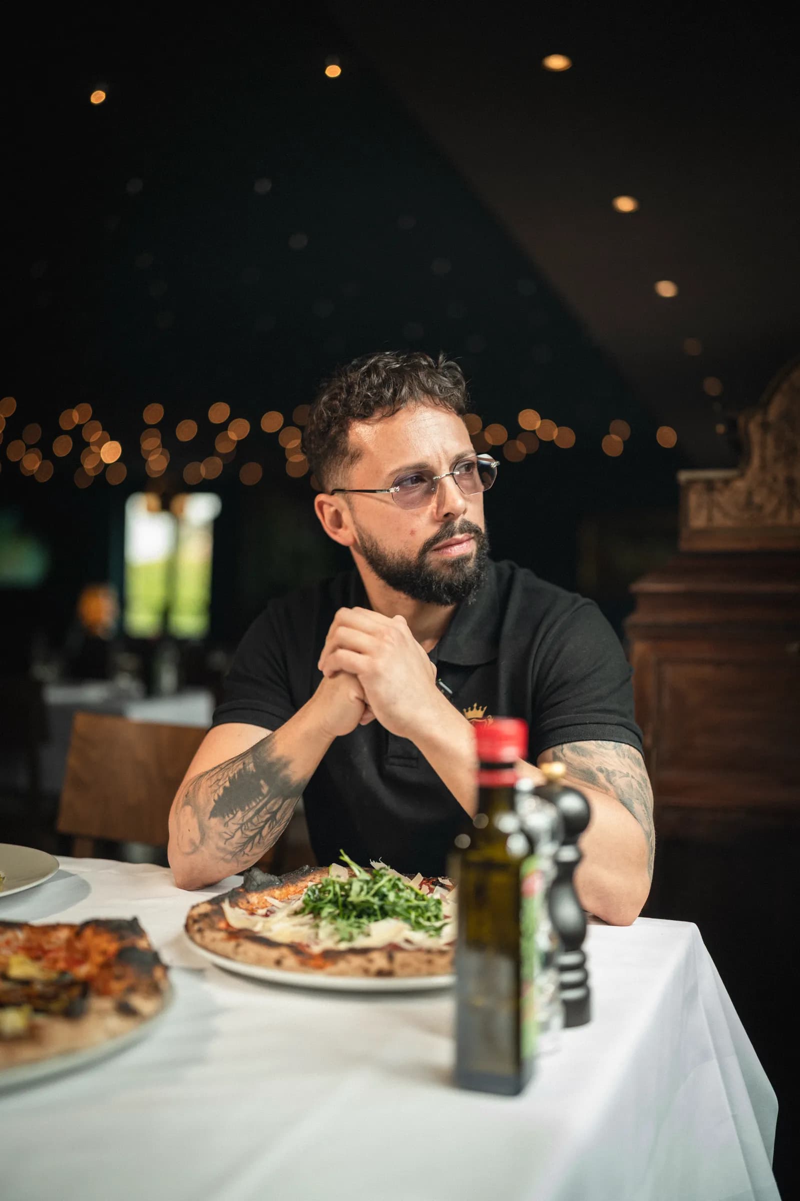 The pizzaiolo seated at a white-clothed table with his pizzas, a bottle of olive oil in the foreground and string lights behind.