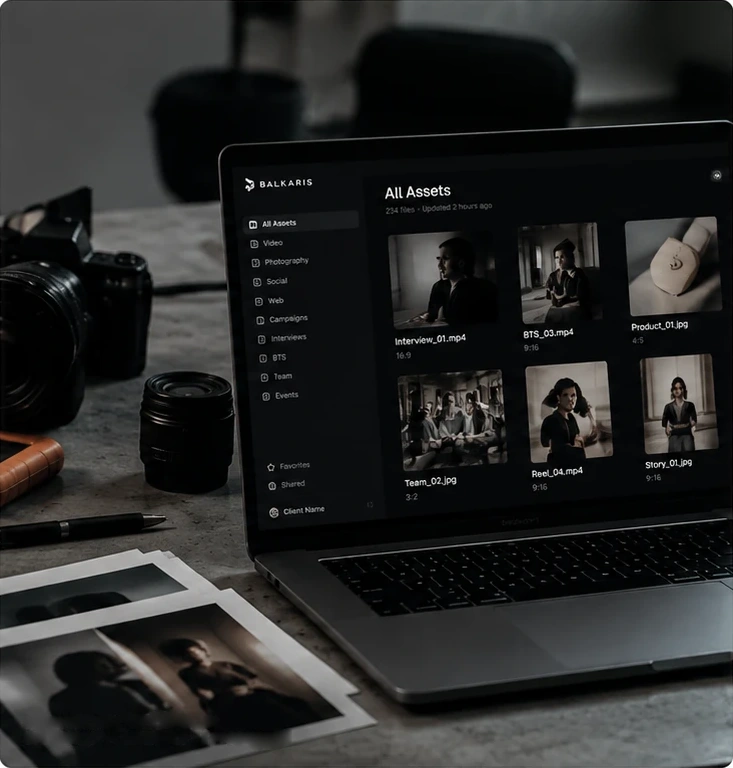 A laptop on a stone desk showing a Balkaris asset library of interviews, behind-the-scenes, product shots, reels and stories, beside a camera, a lens, a pen and a stack of printed photographs.