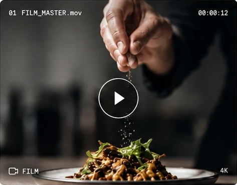 A chef's hand scattering seasoning over a plate of pasta and greens, a play button over the frame.