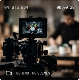 A camera rig on set, its monitor showing the dish being filmed while a chef works behind it.