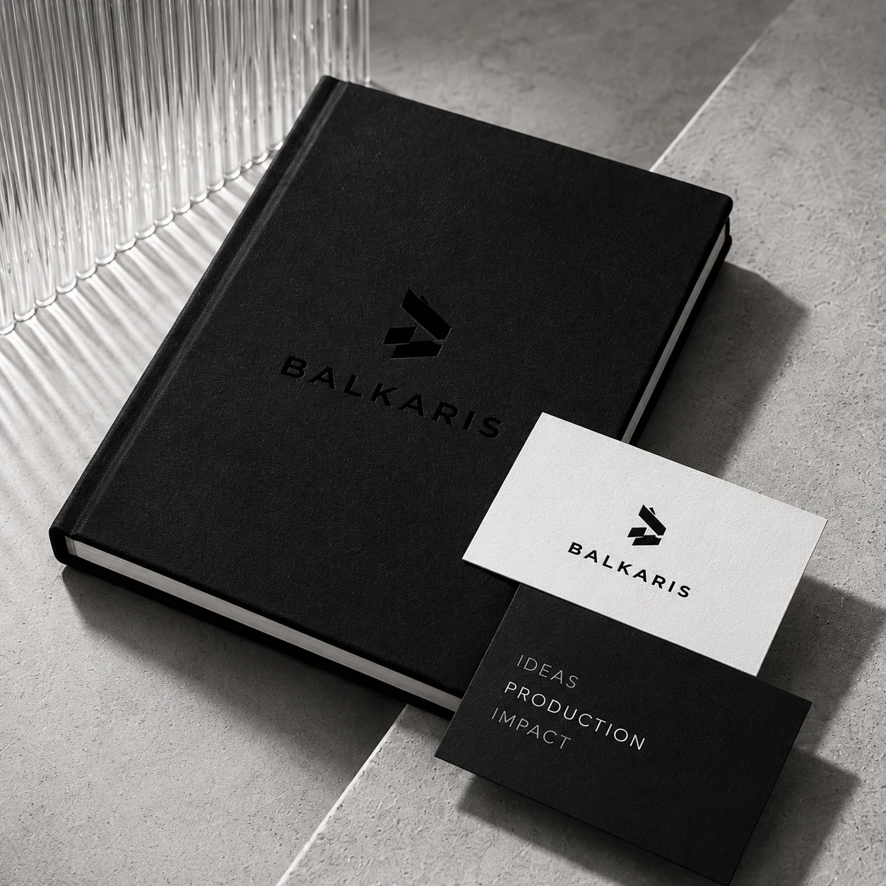 A black clothbound book with the Balkaris mark, a white card and a black card reading Ideas, Production, Impact, on a stone step beside fluted glass