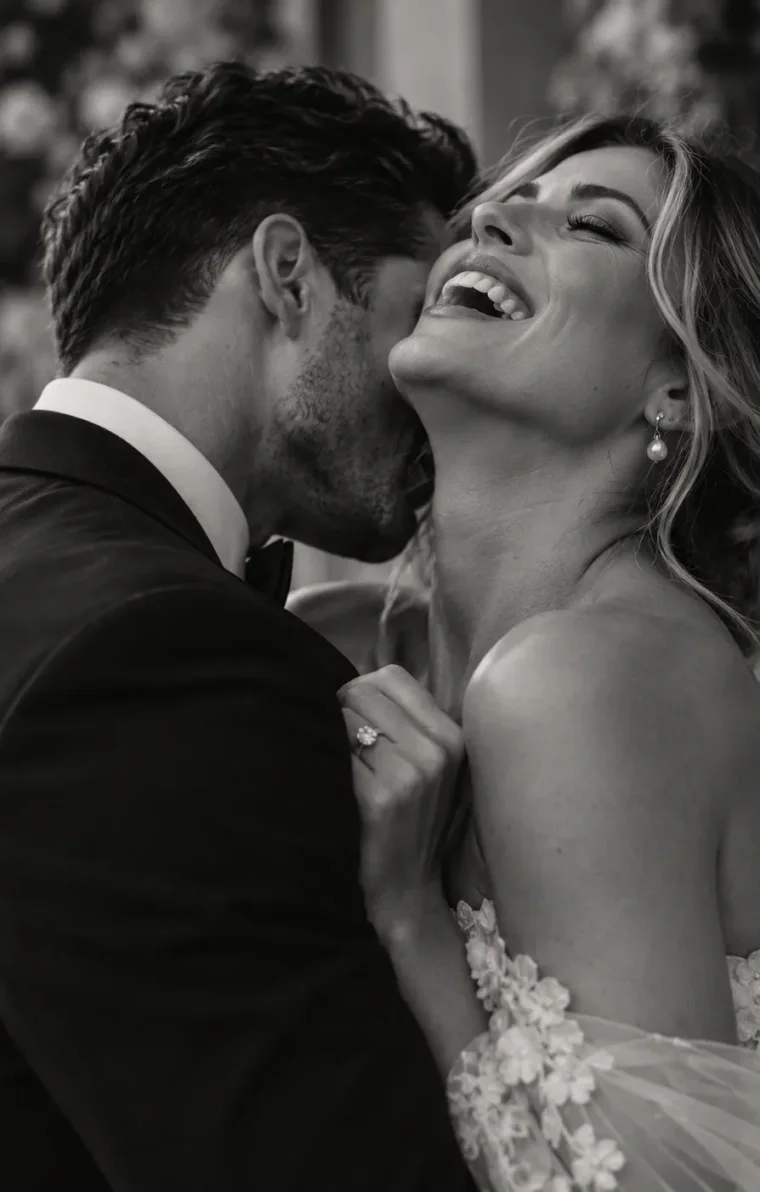 A groom kissing his bride's neck while she laughs, in black and white.
