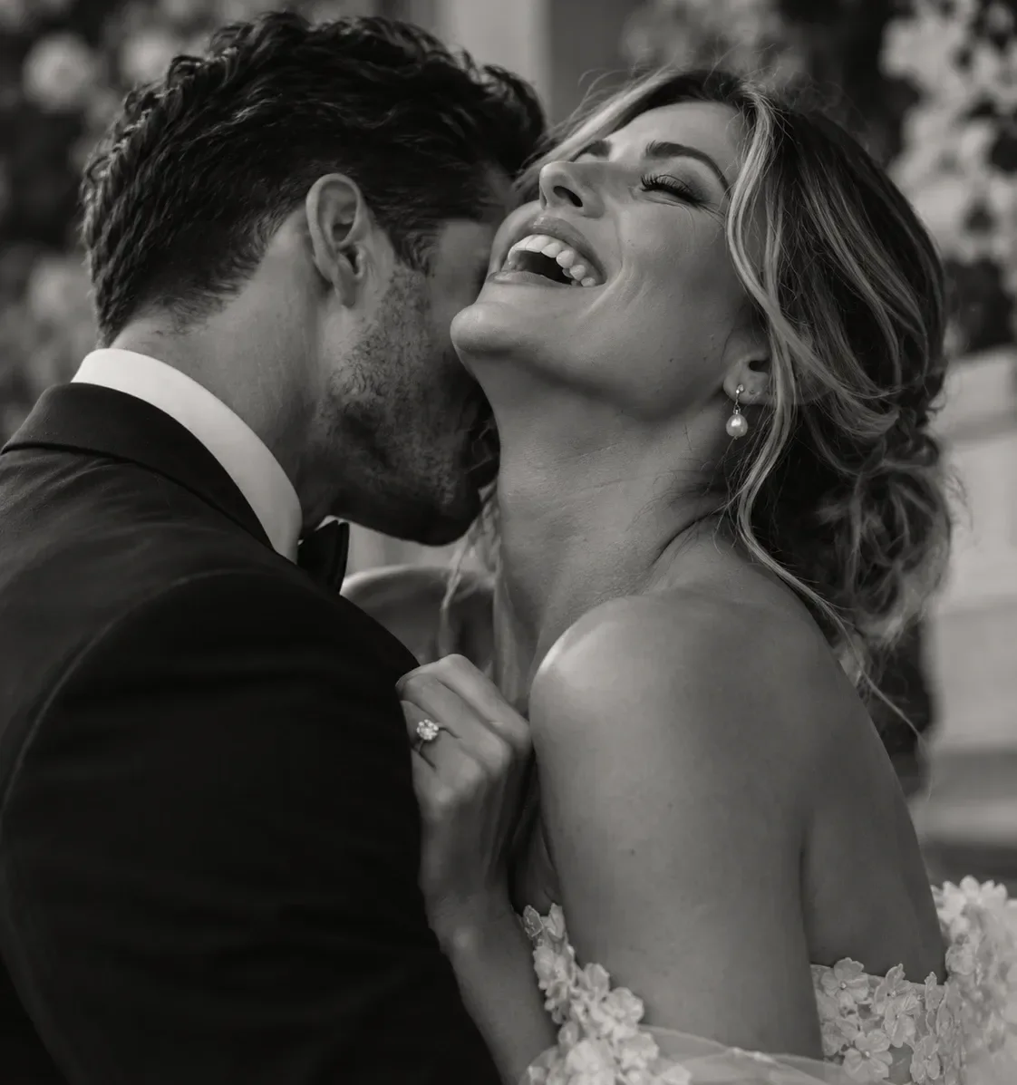 A groom with his face against his bride's neck while she laughs with her head thrown back, in black and white.