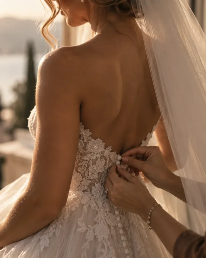 Hands doing up the covered buttons at the back of a lace wedding dress, the veil falling beside them.