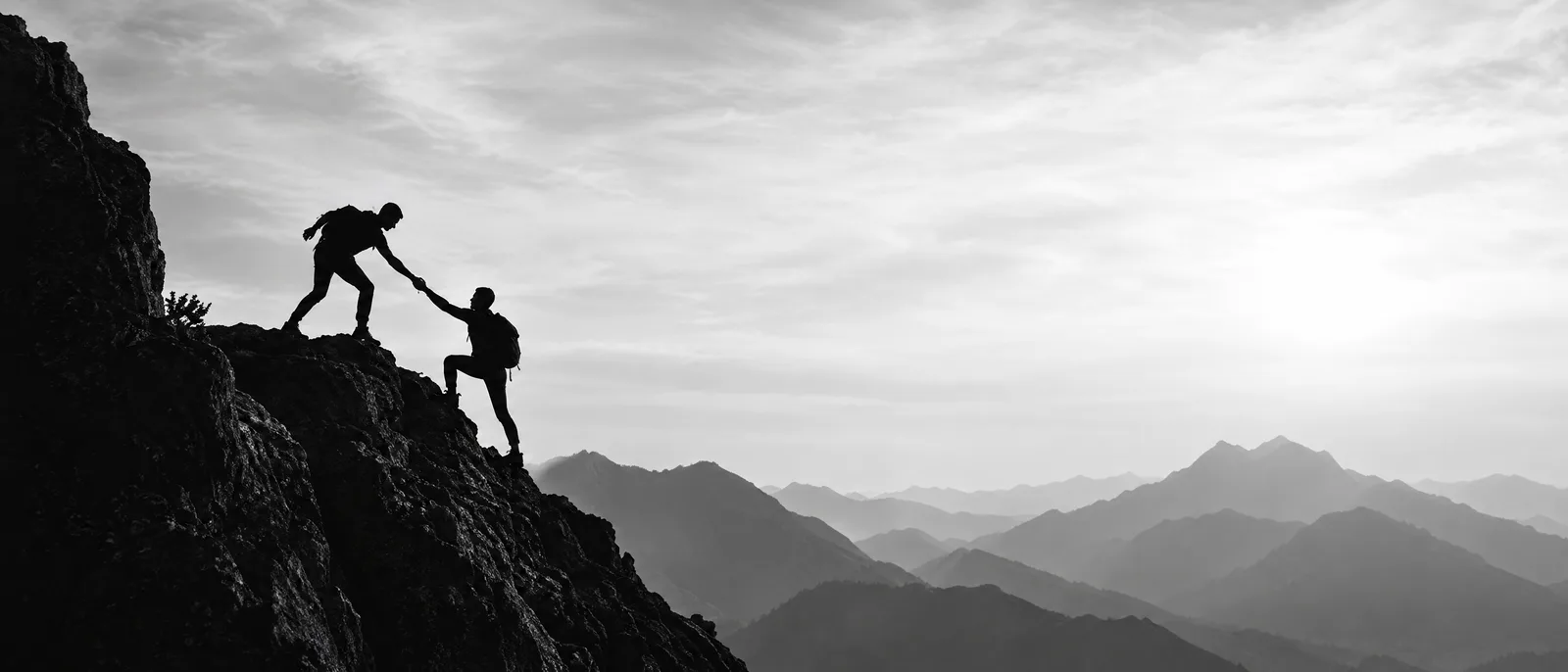Two hikers on a mountain ridge, one reaching down to pull the other up, ranges fading into haze behind them.